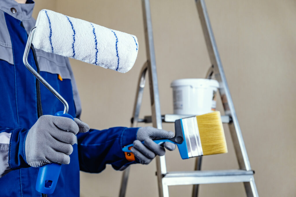 the plaster painter is ready to paint the wall. in the hands of a roller and brush. a stepladder and a bucket of paint in the background.